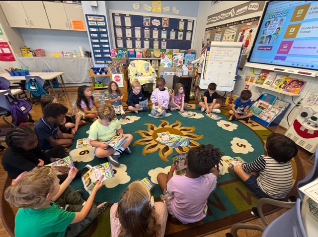 A group of students sit in a circle on a classroom rug and read books together