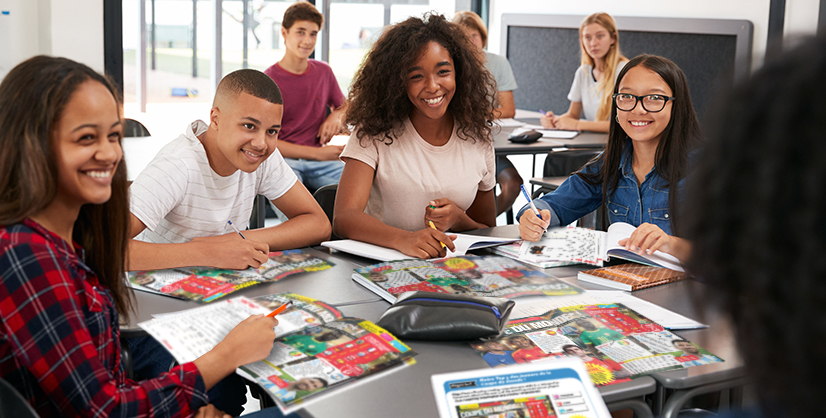 A group of students sit at a classroom table with open books and notebooks