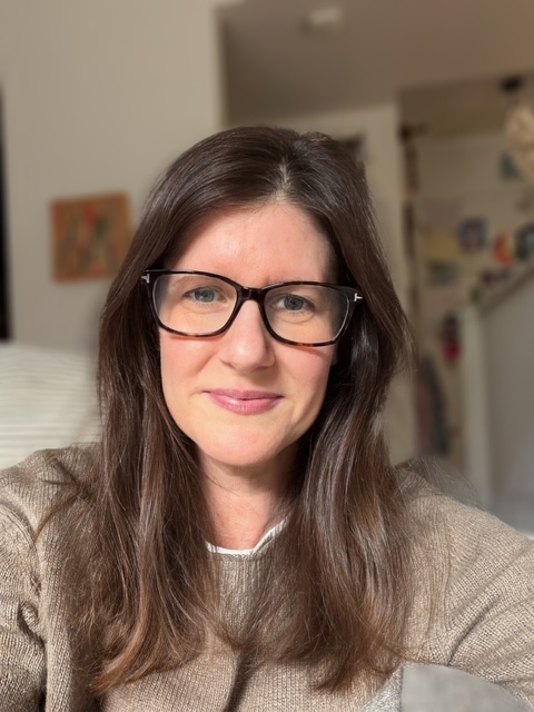 A woman with glasses and long brown hair faces the camera indoors
