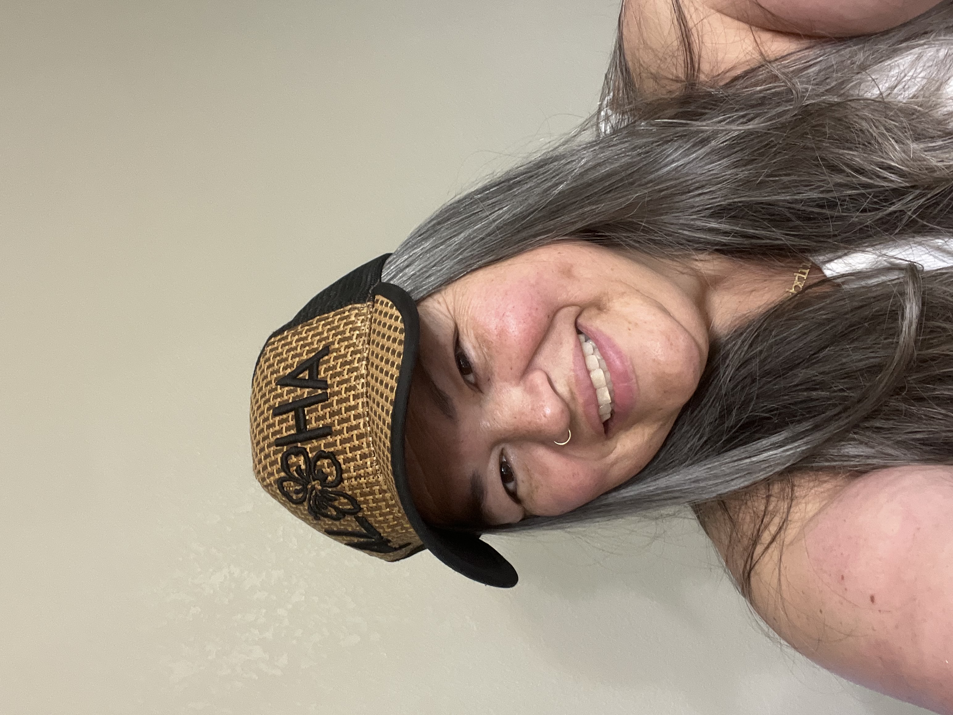 A woman with long gray hair wears a woven snapback cap with embroidered text