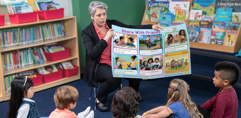 A woman holds up a Share With Friends poster for a group of young children seated on a classroom floor