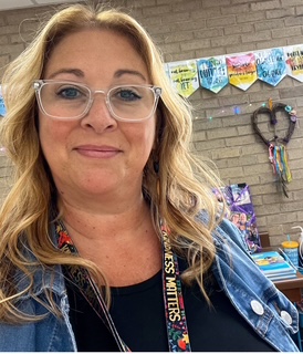 A woman with glasses stands in front of a decorated classroom wall with colorful banners