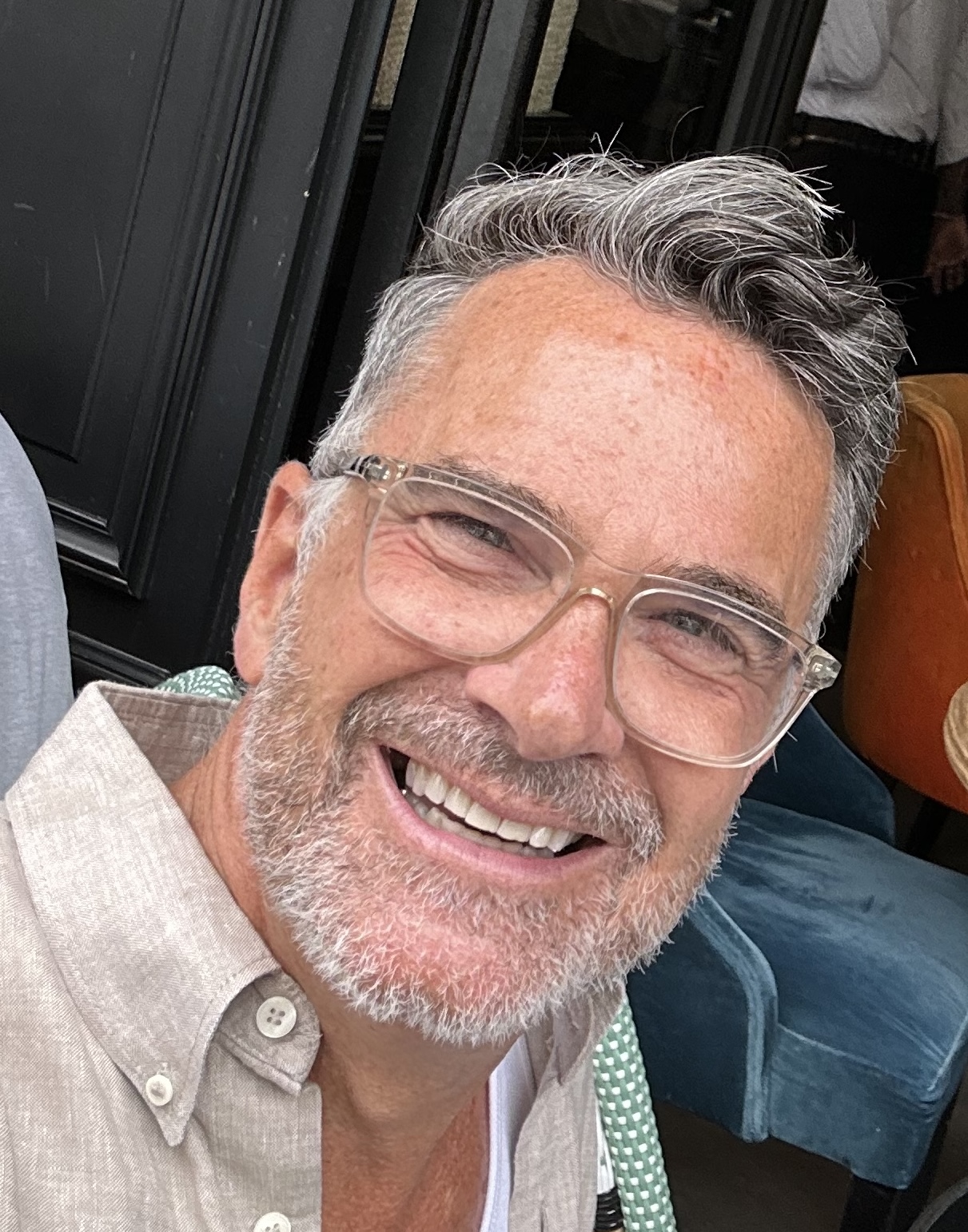 A man with gray hair and clear-framed glasses faces the camera with an open smile
