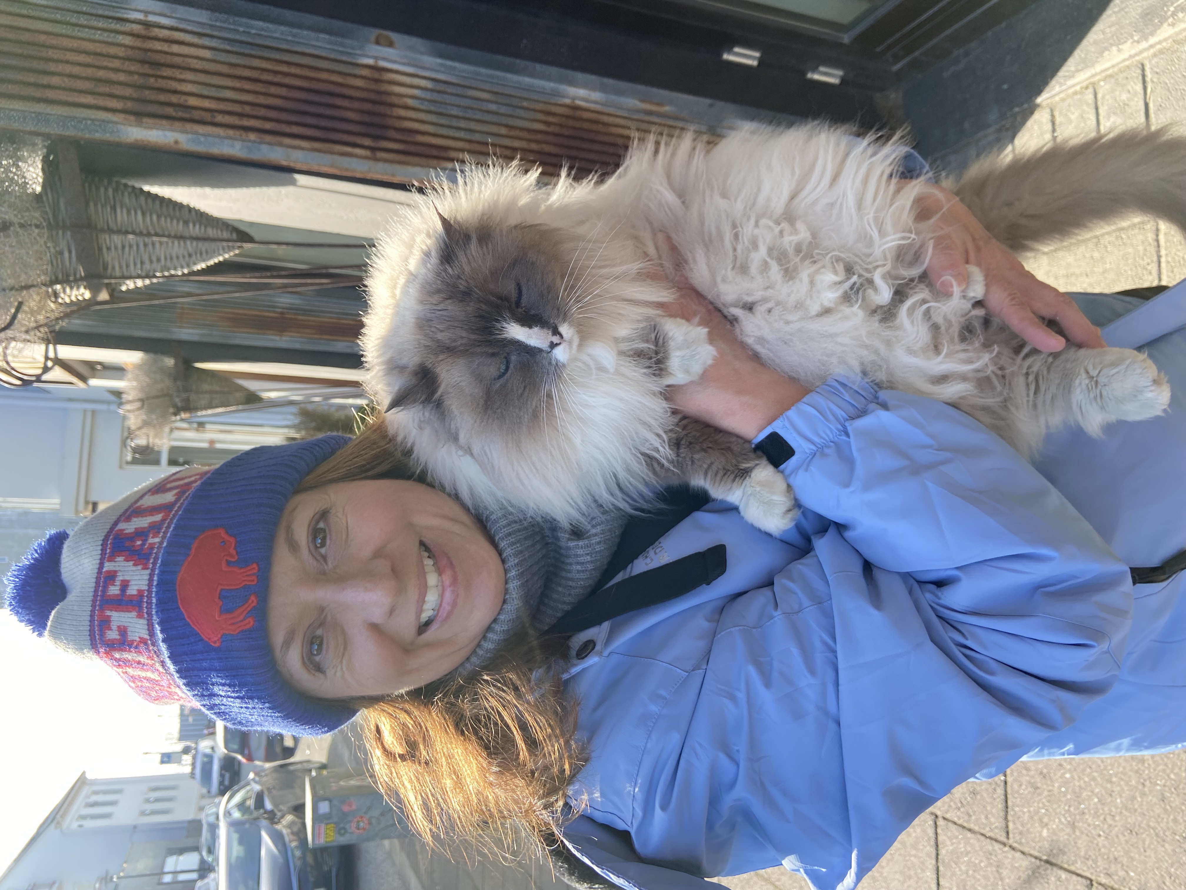 A woman holds a large fluffy cat on a city sidewalk while wearing a Buffalo Bills winter hat