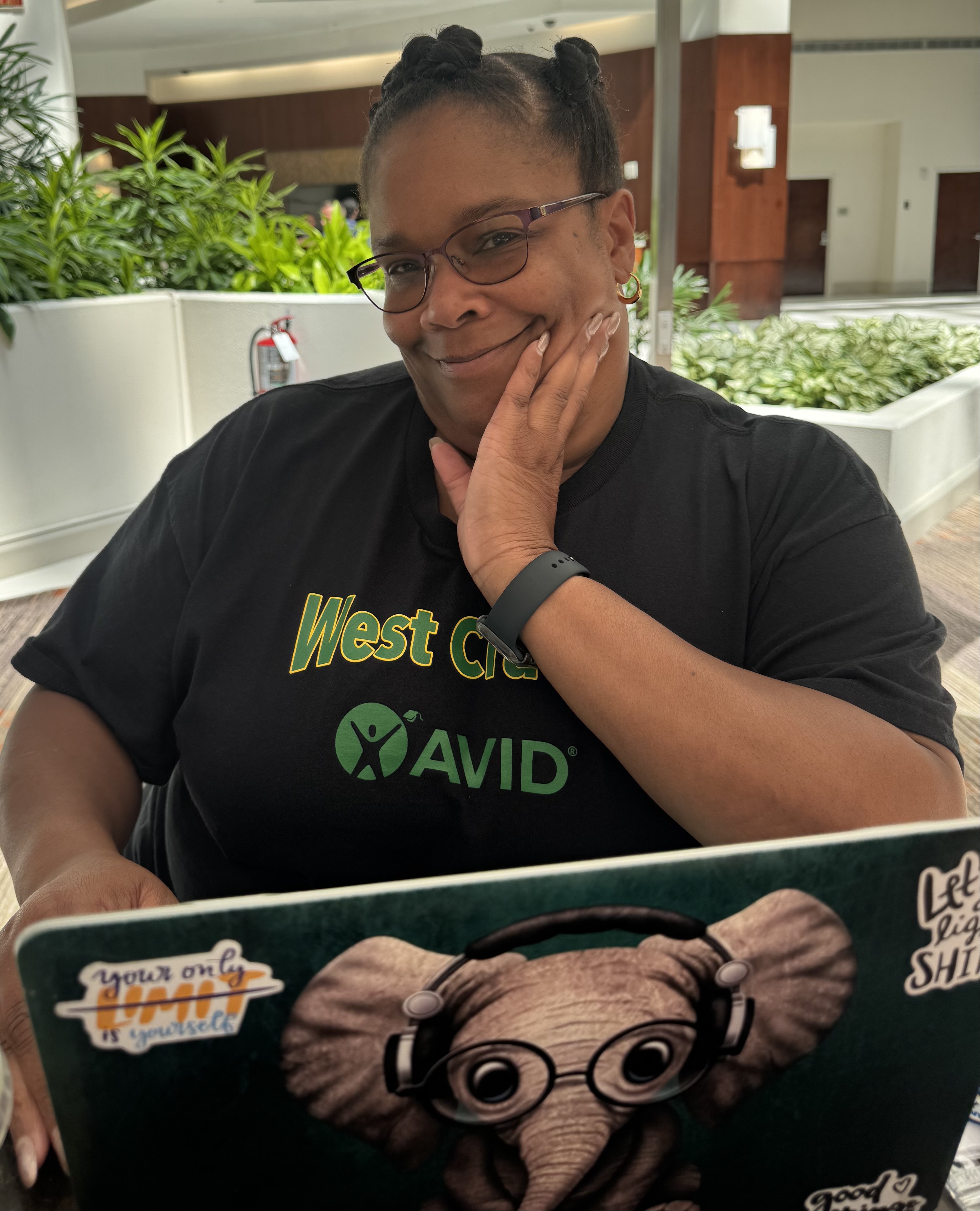 A woman in an Avid shirt rests her chin on her hand behind a sticker-covered laptop