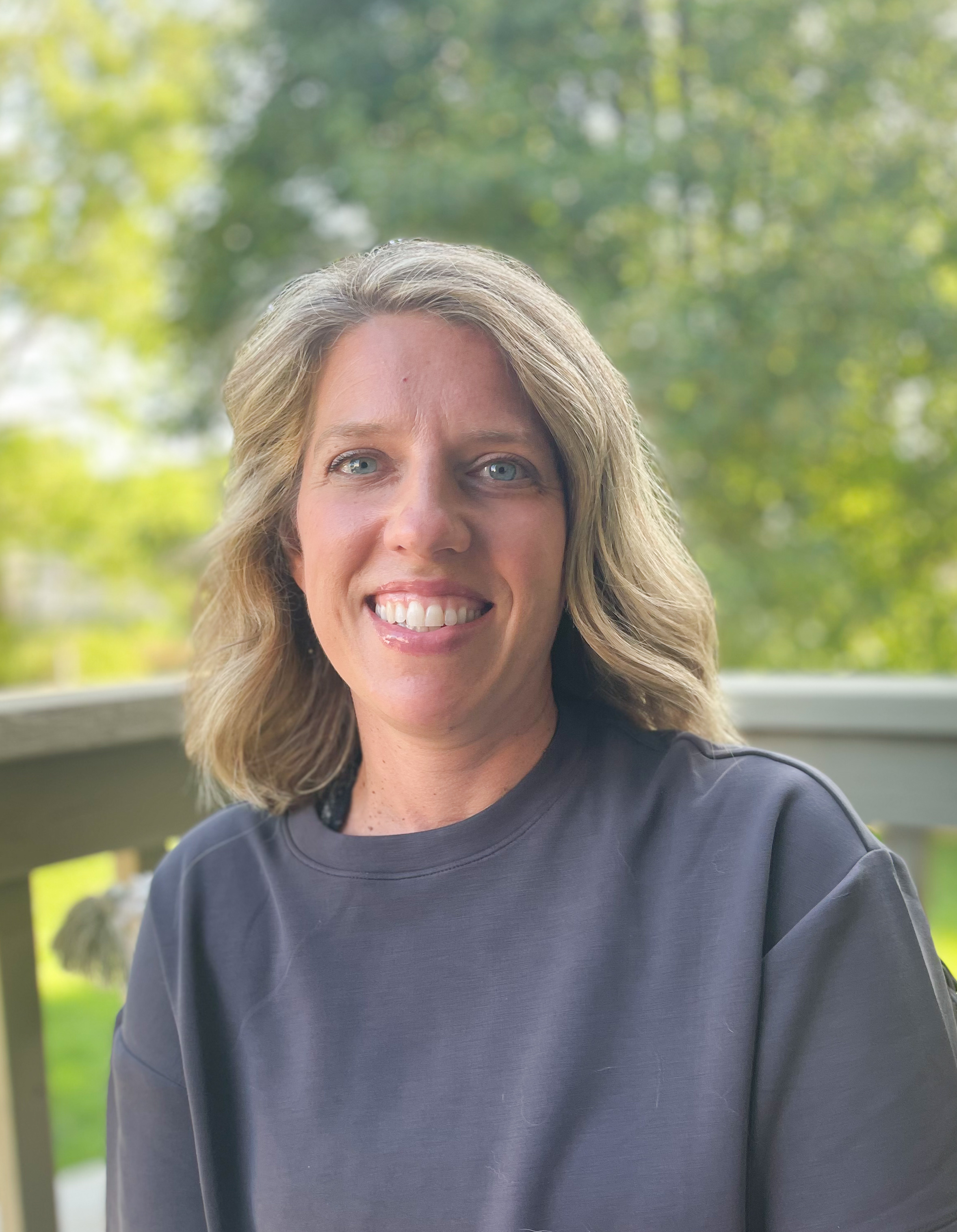 A woman sits outdoors on a deck with trees visible in the background