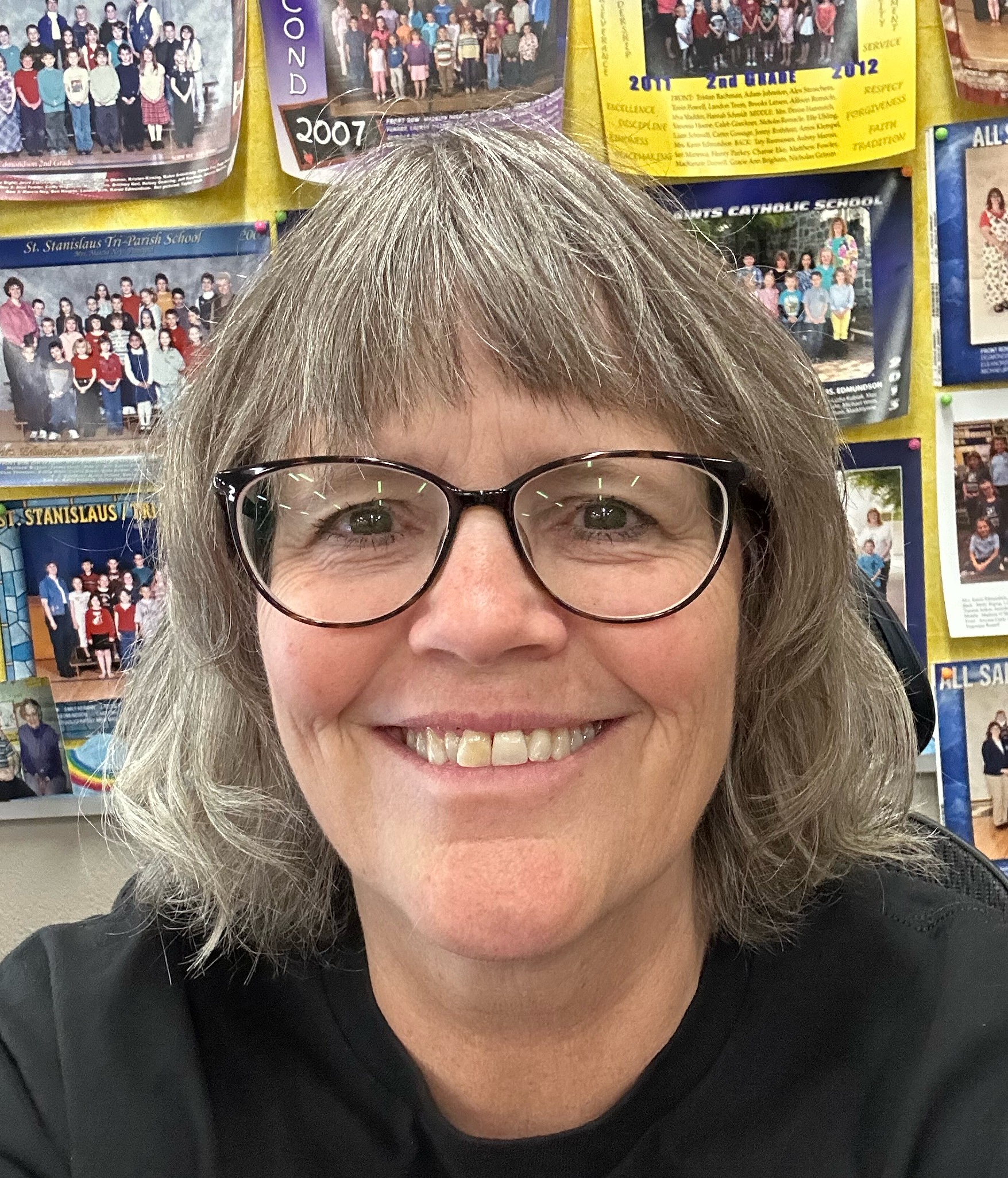 A woman with glasses poses in front of a wall covered with school class photos