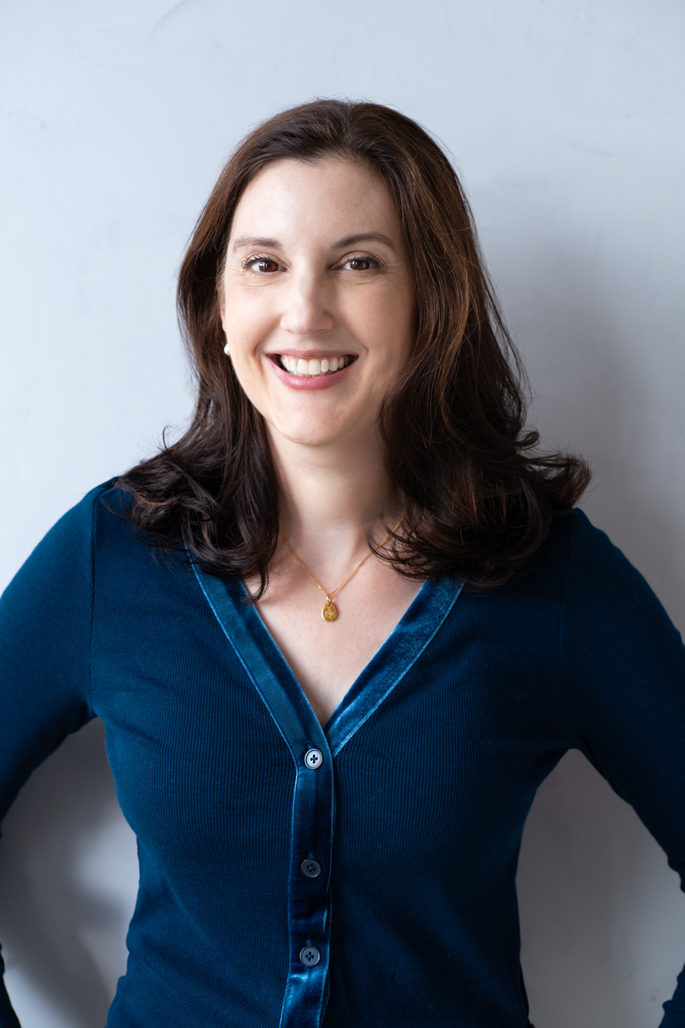 A woman stands with hands on hips and faces the camera against a plain background