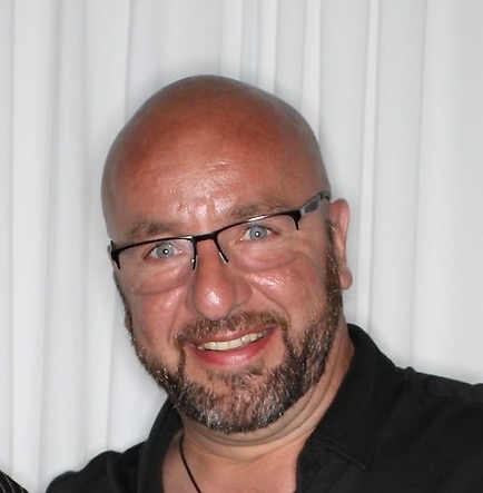 A man with glasses and a beard faces the camera with a wide smile