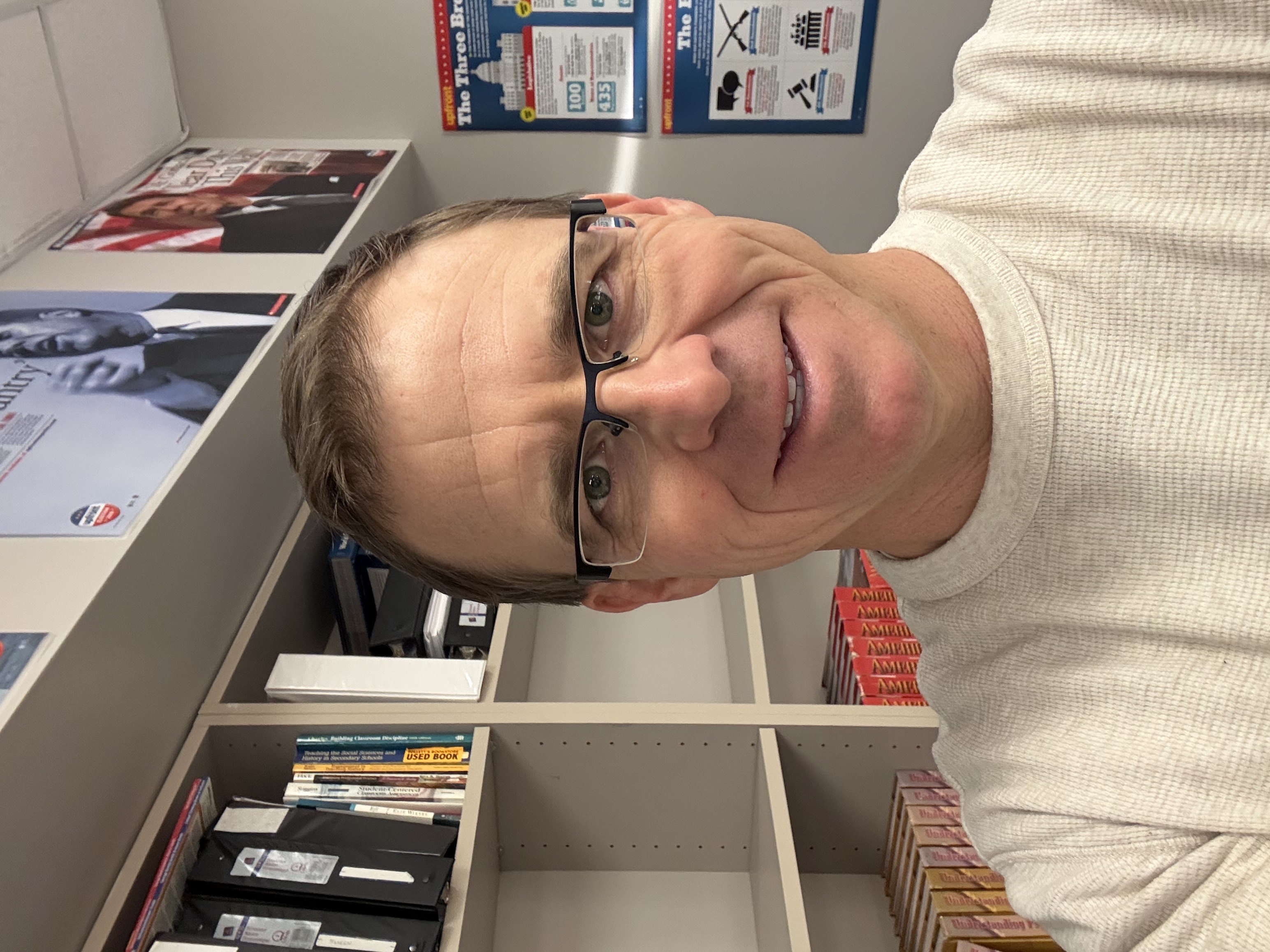 A man with glasses stands in front of a bookshelf and educational posters about government