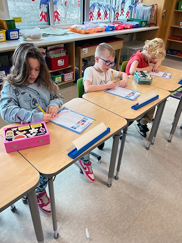 Three students sit at individual desks in a classroom and write on worksheets with pencils