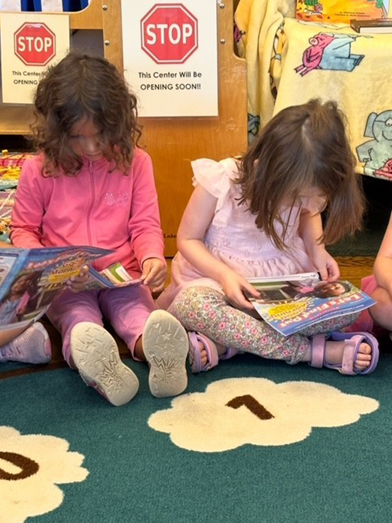 Two girls sit on a classroom floor and read magazines together